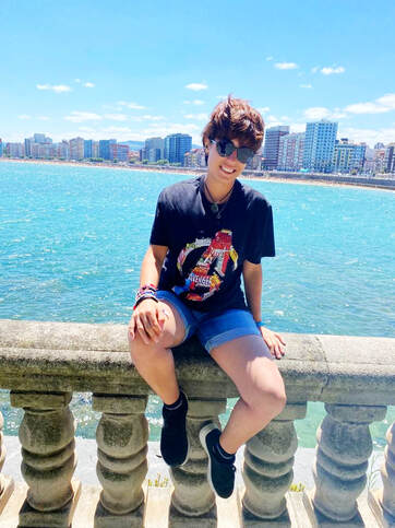 Foto en la que estoy sobre un muro delante de la playa de San Lorenzo. Se ve la primera línea de edificios de Gijón. Llevo una camiseta negra con el logo de los Vengadores y gafas de sol.